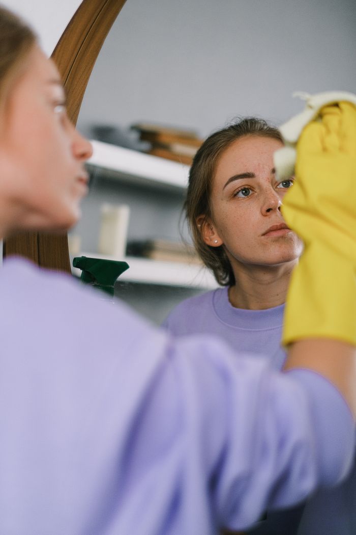 hero-02 Crop female in purple sweatshirt and latex gloves washing mirror in light apartment in daytime