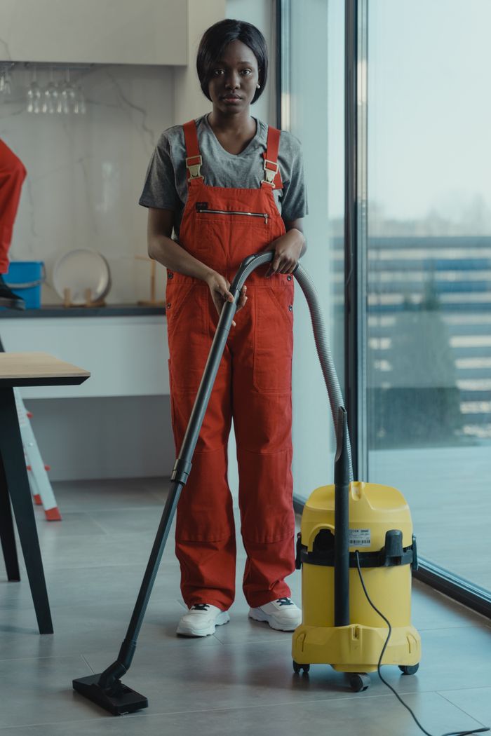 hero-01 An adult woman in red coveralls operates a vacuum cleaner in a modern indoor setting, focusing on cleaning tasks.